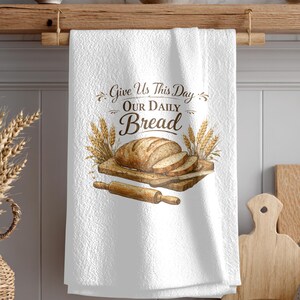 Puede incluir: Paño de cocina blanco con el texto "Give Us This Day Our Daily Bread" y una ilustración con temática de pan. La ilustración presenta una hogaza de pan, pan en rodajas, trigo, una tabla de cortar de madera y un rodillo.