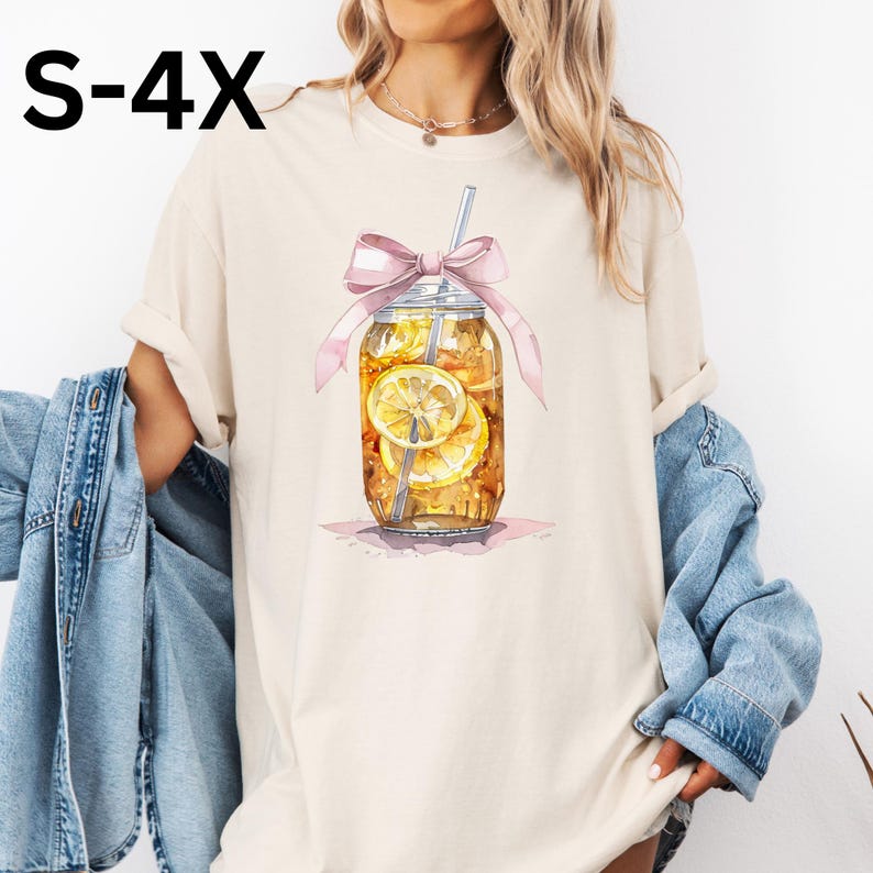 Peut inclure: Un t-shirt beige avec une illustration aquarelle d'un bocal en verre rempli de th&eacute; glac&eacute; et de tranches de citron. Le bocal est attach&eacute; avec un ruban rose et a une paille qui sort du haut.