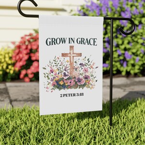 Op de afbeelding: Een witte tuin vlag met een aquarel ontwerp van een houten kruis omringd door roze, paarse en gele bloemen. De tekst "GROW IN GRACE" staat boven het kruis en "2 PETER 3:18" staat onder het kruis.