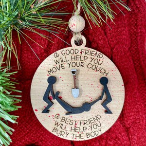 May include: A round wooden Christmas ornament with red paint splatters. It features a black stick figure illustration of two people helping to bury a third person with a shovel. The text reads "A good friend will help you move your couch. A best friend will help you bury the body."