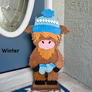 May include: Wooden Highland cow decoration wearing a blue winter hat and mittens with snowflake designs. The cow has a light brown face and body, and the word "Winter" is printed on the door behind it.