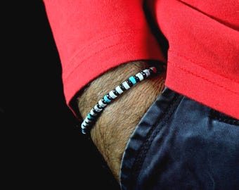 May include: A beaded bracelet with black, white, and turquoise beads. The bracelet is worn on a wrist, with a red long-sleeve shirt and dark blue pants in the background. The bracelet is a simple, yet stylish accessory.
