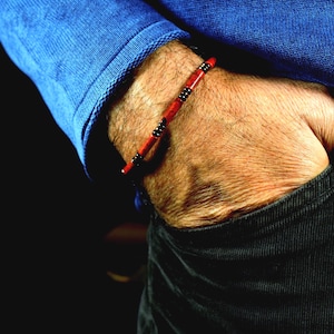Puede incluir: Pulsera de cuentas rojas y negras. La pulsera presenta cuentas rojas intercaladas con cuentas negras y plateadas. La pulsera se lleva en la muñeca, con una camisa azul de manga larga y pantalones negros en el fondo.