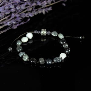 Pode incluir: Uma pulseira de contas com pedras pretas, cinzentas e brancas. A pulseira tem um fecho de nó deslizante e uma conta de destaque prateada. Fundo preto, com um ramo de flores roxas.