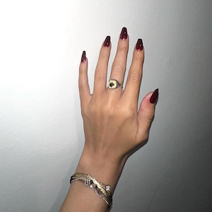 Op de afbeelding: Een hand met lange, bordeauxrode nagels, met een gouden ring met een donkere middensteen en een gouden armband. De hand bevindt zich tegen een lichtgrijze achtergrond.