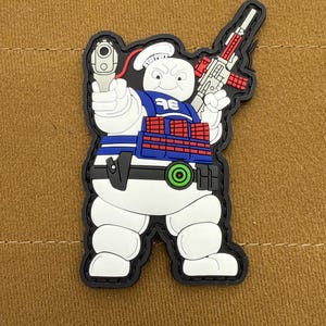 May include: A white marshmallow man character with a blue vest and red ammunition belt, holding a gun in one hand and a rifle in the other. The character is wearing a white chef's hat with the words "Stay Puft" written on it.