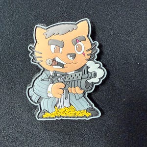 May include: A cartoon-style pin featuring a cat character in a gray pinstripe suit, holding a machine gun with smoke. The cat has a cigar in its mouth and is surrounded by yellow objects. The pin has a black border.