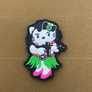 May include: A black and white cartoon patch of a Hello Kitty character wearing a green grass skirt and holding a black gun. The character has a pink bow and a green target on her head. The patch has a pink and green flower design.