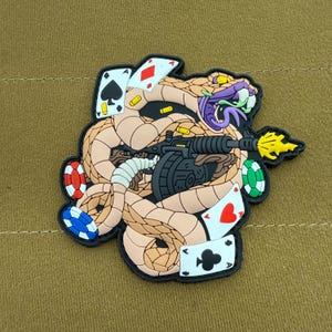 May include: A brown snake with a gun and playing cards wrapped around it. The snake is holding a gun with a yellow flame coming out of the barrel. The snake is also holding playing cards, including a red ace of hearts, a black ace of spades, and a black jack of spades. The snake is surrounded by poker chips.