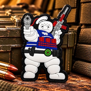 May include: A rubberized morale patch featuring a white Stay Puft marshmallow character wearing a blue tactical vest with red magazine pouches, holding a gray handgun in one hand and a gray rifle in the other, with a black belt and green target icon.