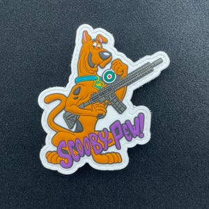 May include: A cartoon patch featuring Scooby-Doo holding a rifle. The orange dog is wearing a blue collar and a green tag. The text "SCOOBY-PEW!" is in purple. The patch has a white border.