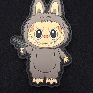 May include: A gray cartoon character patch with a rabbit-like head, holding a black handgun. The character has large eyes, a toothy grin, and orange cheeks. The patch has a black border.