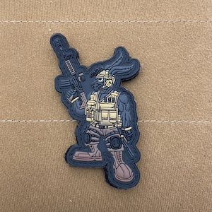 May include: Black and tan embroidered patch of a rabbit wearing tactical gear and holding a rifle. The rabbit is wearing a helmet, a tactical vest, and gloves. The patch is about 2 inches tall.
