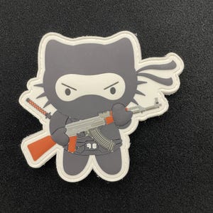 May include: A white and gray patch with a cartoon image of a Hello Kitty-like character wearing a black ninja mask and holding a red and silver AK-47 rifle.