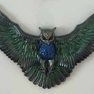 May include: A black, green, and blue owl figurine with red eyes. The owl is in flight with its wings spread wide. The figurine is made of a shiny, metallic material.