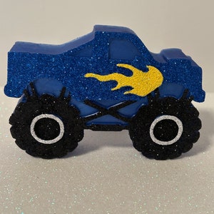 Pu&ograve; includere: Topper per torta a forma di monster truck blu con pneumatici neri e un design a fiamma gialla.