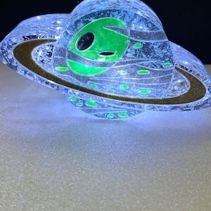 May include: A clear glass sphere with a green alien face inside, surrounded by a gold ring, resembling a planet with rings. The sphere is illuminated from within, creating a glowing effect.