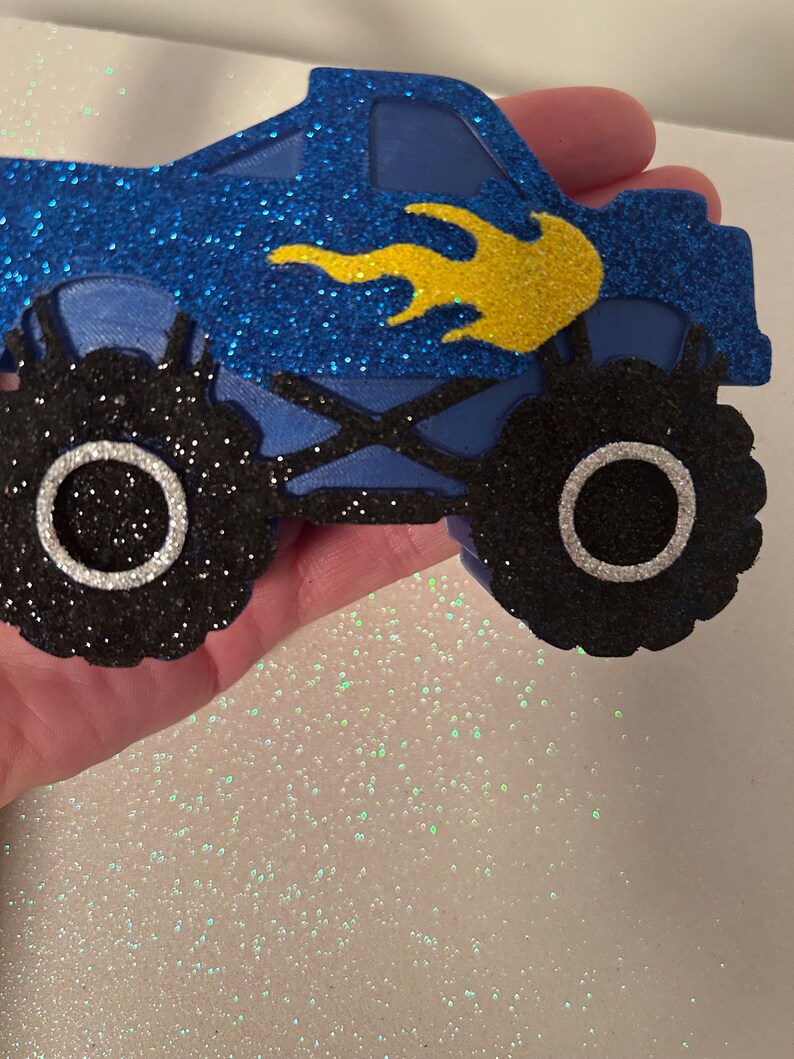 Pu&ograve; includere: Monster truck blu e nero con glitter e fiamme gialle. Il camion ha grandi ruote nere con cerchi argentati.