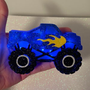 Pu&ograve; includere: Un monster truck blu con glitter, fiamme gialle e pneumatici neri. Il camion ha un cerchio bianco attorno a ogni pneumatico. Il camion &egrave; illuminato da luci blu.