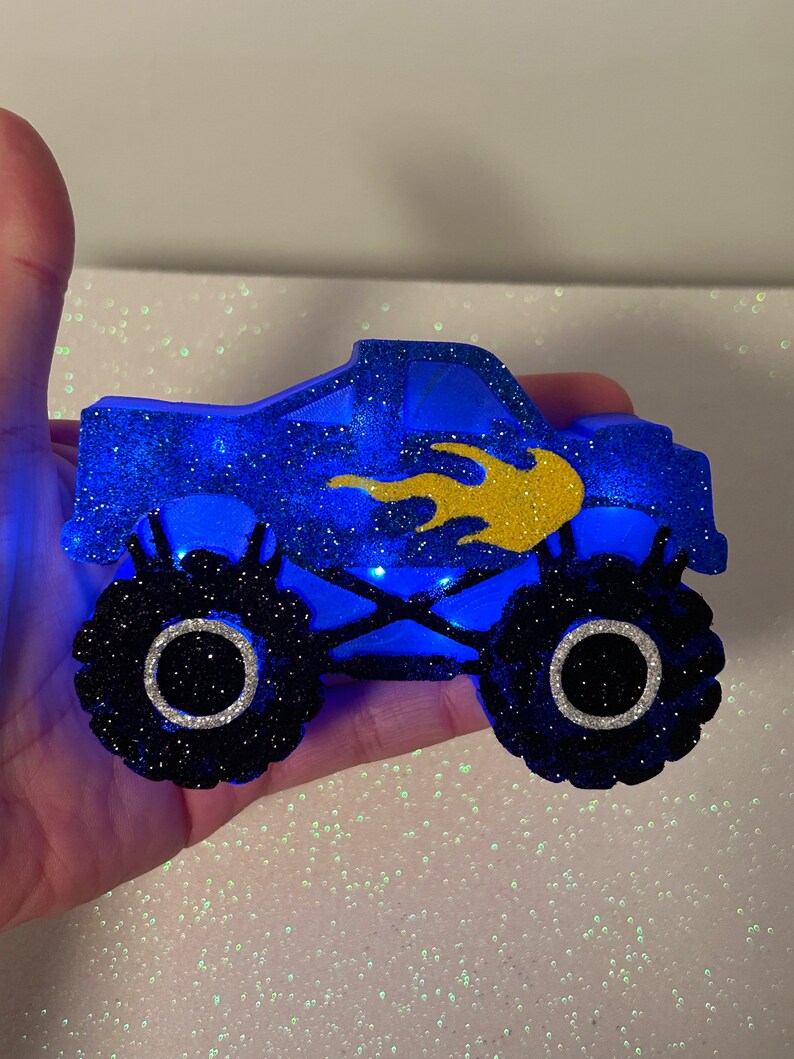 Pu&ograve; includere: Un monster truck blu con glitter, fiamme gialle e pneumatici neri. Il camion ha un cerchio bianco attorno a ogni pneumatico. Il camion &egrave; illuminato da luci blu.
