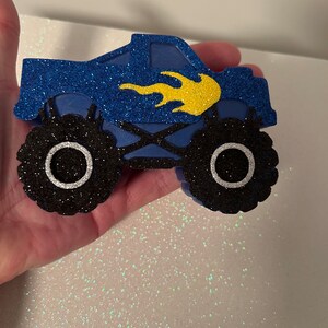 Pu&ograve; includere: Taglio di camion mostro blu e nero con un design a fiamma gialla. Il camion ha grandi ruote nere con cerchi bianchi.