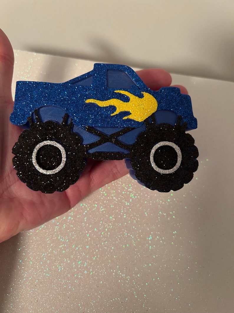 Pu&ograve; includere: Taglio di camion mostro blu e nero con un design a fiamma gialla. Il camion ha grandi ruote nere con cerchi bianchi.