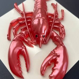May include: A red plastic lobster figurine with a glossy finish. The lobster is posed with its claws raised and its antennae extended.