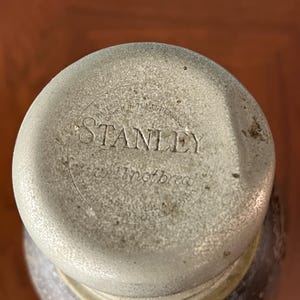 May include: Close-up of a Stanley thermos lid. The lid is a light gray color with the word "STANLEY" embossed in the center. The text "will not break" is also visible. The lid is slightly worn, suggesting age.