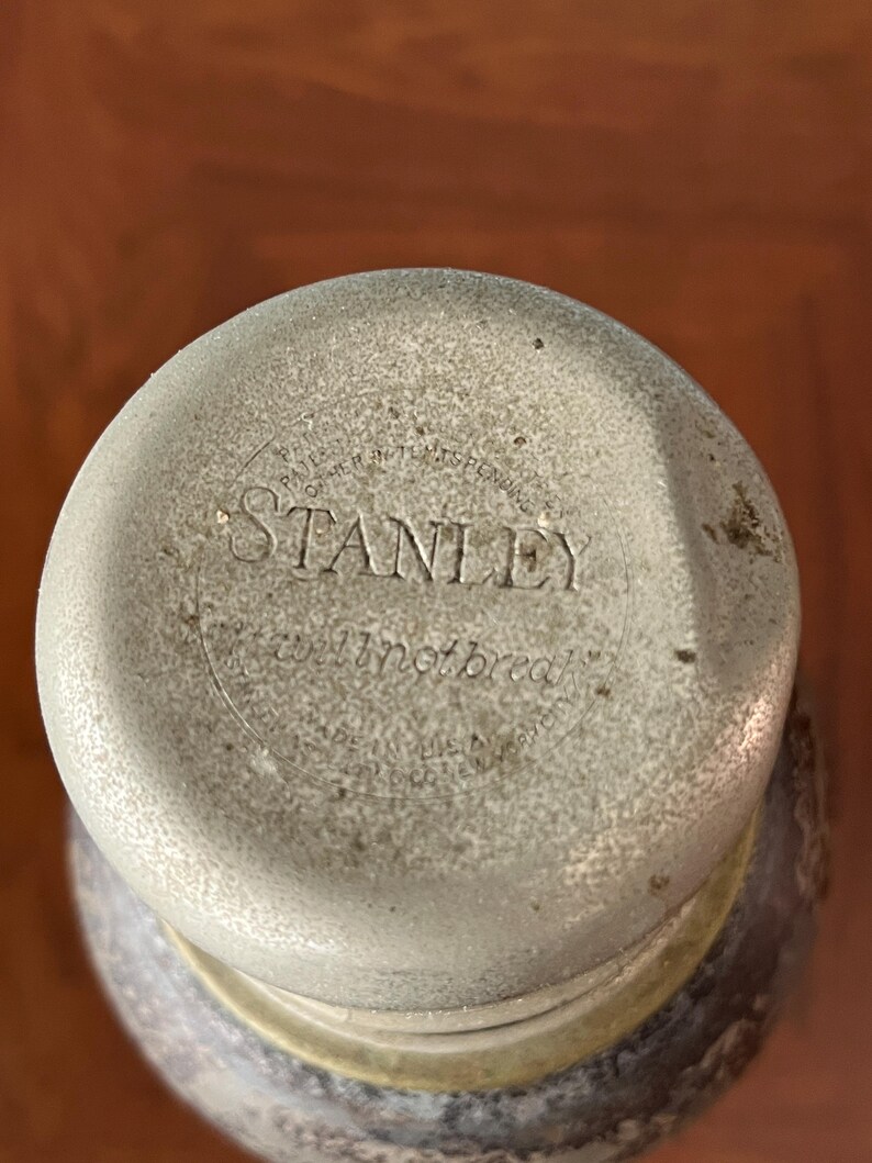 May include: Close-up of a Stanley thermos lid. The lid is a light gray color with the word "STANLEY" embossed in the center. The text "will not break" is also visible. The lid is slightly worn, suggesting age.