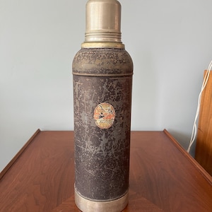 May include: Vintage brown and silver thermos with a metal cap and base. The thermos has a distressed finish and a small circular label. A classic container for hot or cold beverages.