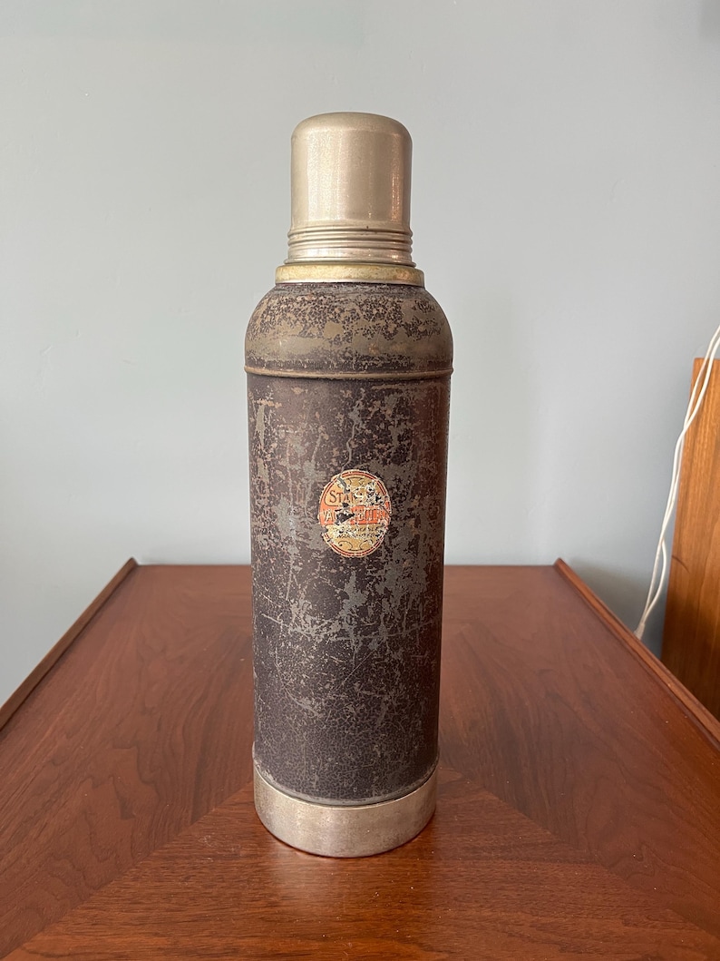 May include: Vintage brown and silver thermos with a metal cap and base. The thermos has a distressed finish and a small circular label. A classic container for hot or cold beverages.