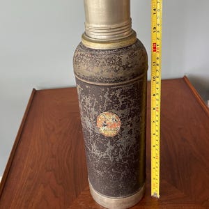 May include: Vintage metal thermos with a distressed finish. The cylindrical body is dark with a worn appearance, and the top and bottom are silver. A label is visible on the side. The thermos is approximately 15 inches tall.