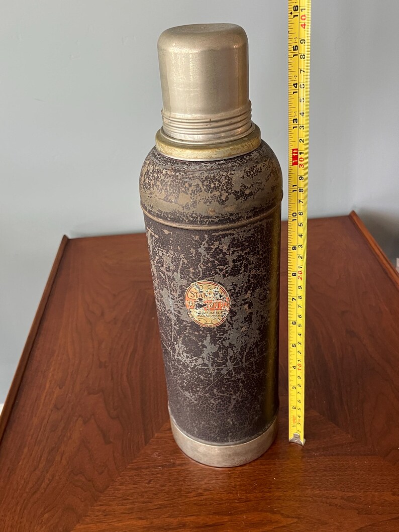 May include: Vintage metal thermos with a distressed finish. The cylindrical body is dark with a worn appearance, and the top and bottom are silver. A label is visible on the side. The thermos is approximately 15 inches tall.
