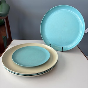 May include: A set of round dinnerware featuring a large upright turquoise plate with a speckled finish, stacked cream-colored plates, and a smaller turquoise plate centered on top. The plates have smooth, flat surfaces and simple circular rims.