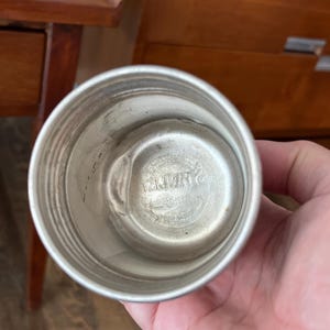 May include: A silver-colored metal cup with a rounded base and a slightly flared rim. The interior has a circular indentation with embossed text. The cup is held in a person's hand.