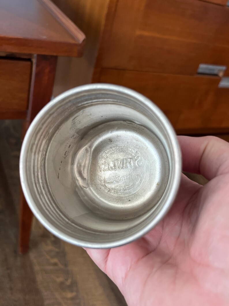 May include: A silver-colored metal cup with a rounded base and a slightly flared rim. The interior has a circular indentation with embossed text. The cup is held in a person's hand.