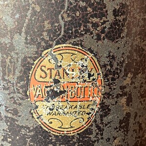 May include: Close-up of a vintage Stanley Vacuum Bottle. The bottle has a distressed, textured surface with a faded label. The label is orange and gold with the words "Stanley Vacuum Bottle" and "Unbreakable Warranted".