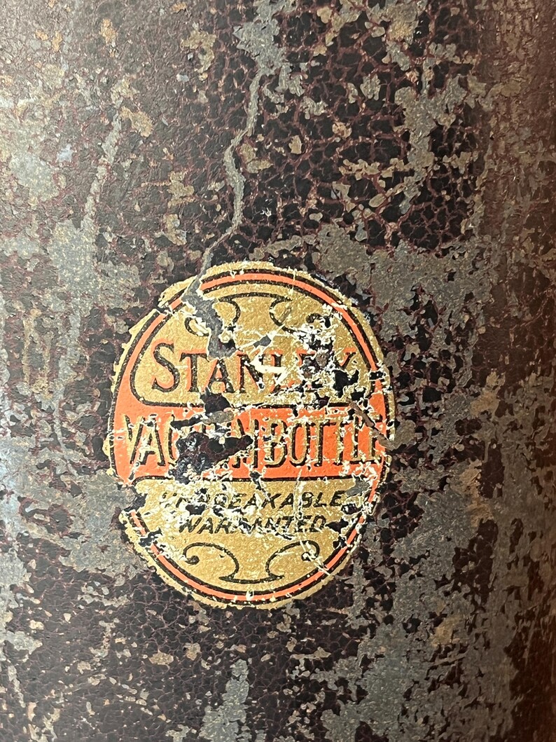 May include: Close-up of a vintage Stanley Vacuum Bottle. The bottle has a distressed, textured surface with a faded label. The label is orange and gold with the words "Stanley Vacuum Bottle" and "Unbreakable Warranted".