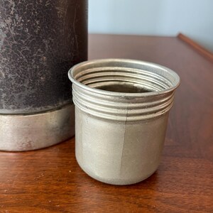 May include: A silver-colored metal cup with a ridged exterior sits next to a dark brown, textured container. The cup has a cylindrical shape with a slightly flared rim. The background is a wooden surface.