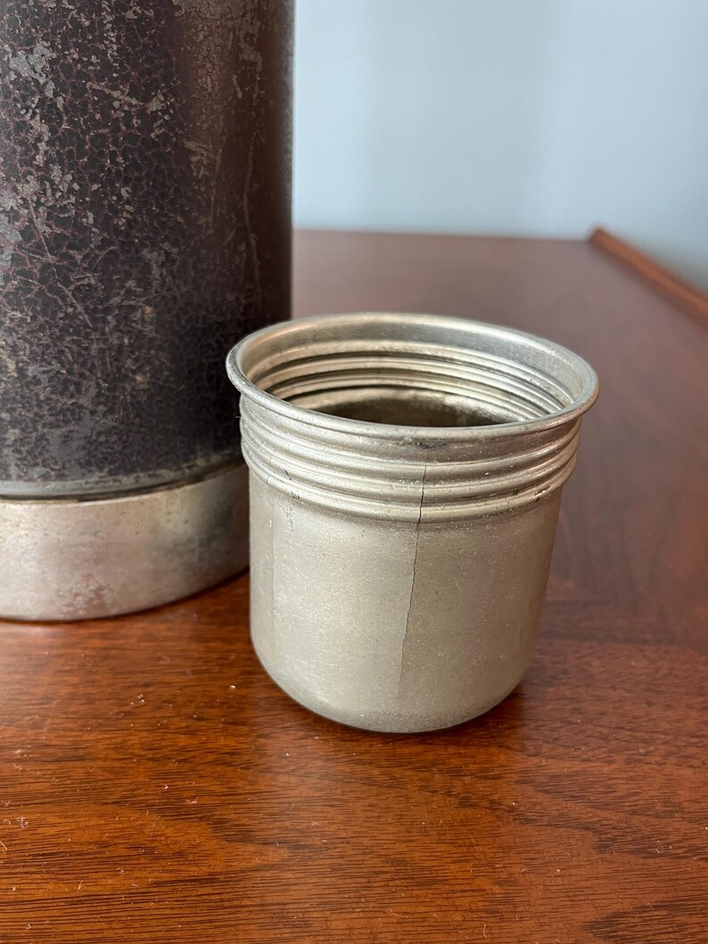 May include: A silver-colored metal cup with a ridged exterior sits next to a dark brown, textured container. The cup has a cylindrical shape with a slightly flared rim. The background is a wooden surface.