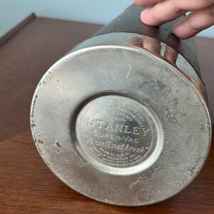May include: A vintage Stanley Super-Vac thermos. The bottom of the thermos is silver-colored metal with the text "STANLEY SUPER-VAC It will not break". The body is black with a silver band around the top.