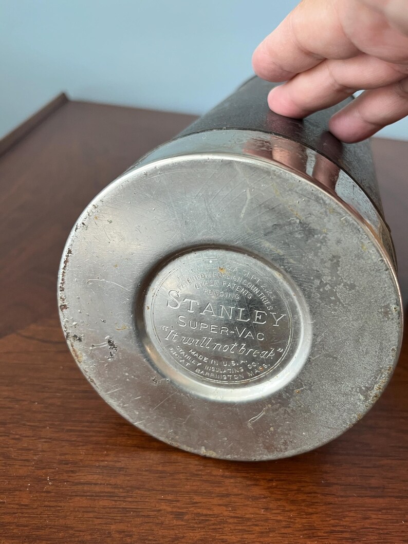 May include: A vintage Stanley Super-Vac thermos. The bottom of the thermos is silver-colored metal with the text "STANLEY SUPER-VAC It will not break". The body is black with a silver band around the top.