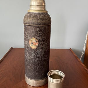 May include: An antique metal thermos with a silver cap and base, showing signs of wear. A small silver cup sits beside the thermos. The thermos has a faded label. The overall color is dark brown with silver accents.