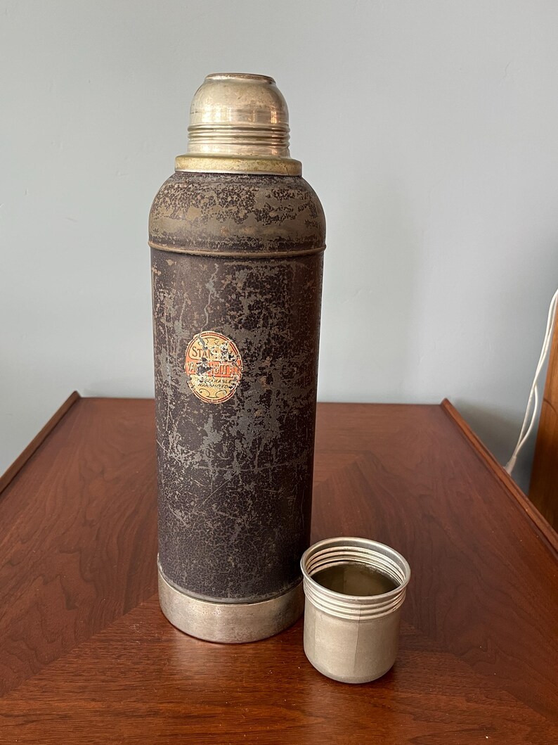 May include: An antique metal thermos with a silver cap and base, showing signs of wear. A small silver cup sits beside the thermos. The thermos has a faded label. The overall color is dark brown with silver accents.