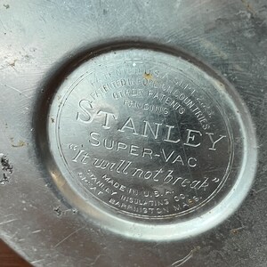 May include: Close-up of a vintage Stanley Super-Vac thermos base. The silver-colored metal base features embossed text including "Stanley Super-Vac" and "It will not break." Made in U.S.A.