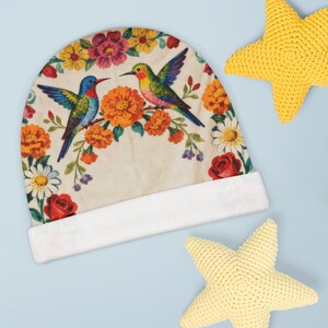 May include: A beanie hat featuring a cream-colored background with a vibrant floral pattern of red roses, orange marigolds, and white daisies. Two hummingbirds, one blue and one multicolored, are positioned among the flowers on the fabric.