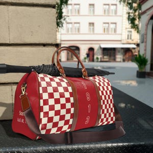 May include: Red and white checkered duffel bag with brown leather handles and trim. The bag has the number "1913" printed vertically on the side. A black umbrella rests on top of the bag. The bag has a brown shoulder strap.