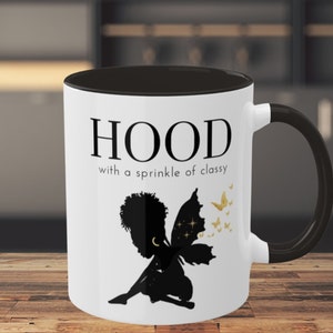 May include: White ceramic mug with a black rim and handle. The mug has a black silhouette of a fairy with wings and a crescent moon on its head. The text "HOOD with a sprinkle of classy" is printed in black on the mug.