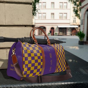 May include: A purple and gold checked duffel bag with brown leather handles and trim. The bag has a shoulder strap and the number "1911" is printed on the side. A black umbrella rests on top of the bag.
