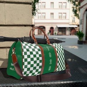 Puede incluir: Una bolsa de deporte a cuadros verdes y blancos con adornos, asas y correa de hombro de aspecto de cuero marrón. La bolsa presenta el texto vertical blanco 1929 y el texto Sorority Ashley L. en el lateral, con un paraguas negro encima.